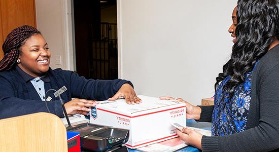 Person drops off 优先邮件 package to 美国邮政总局 retail associate at a Post Office counter.